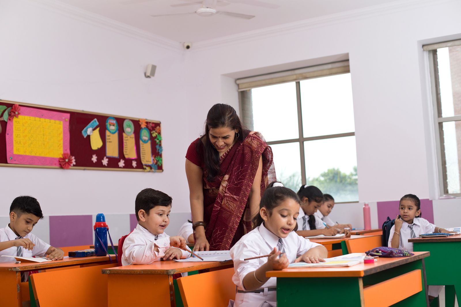 A teacher in a saree guiding young children at their desks in a bright classroom