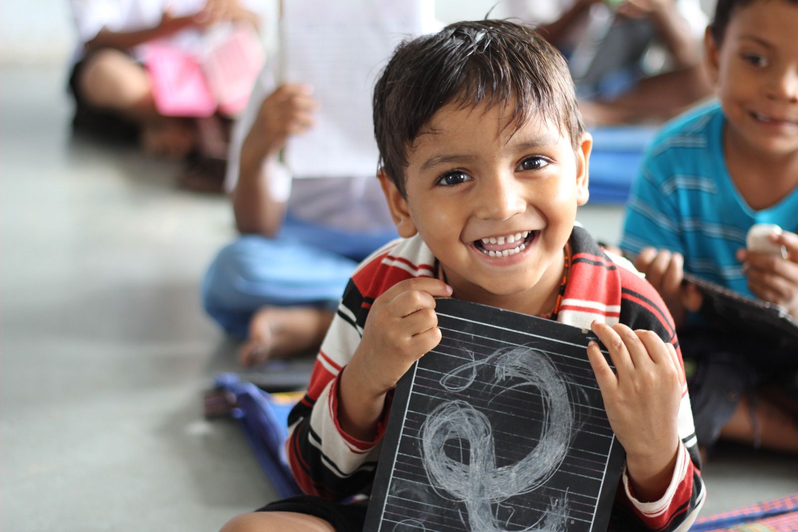 A young boy holding up his slate and smiling on the classroom floor