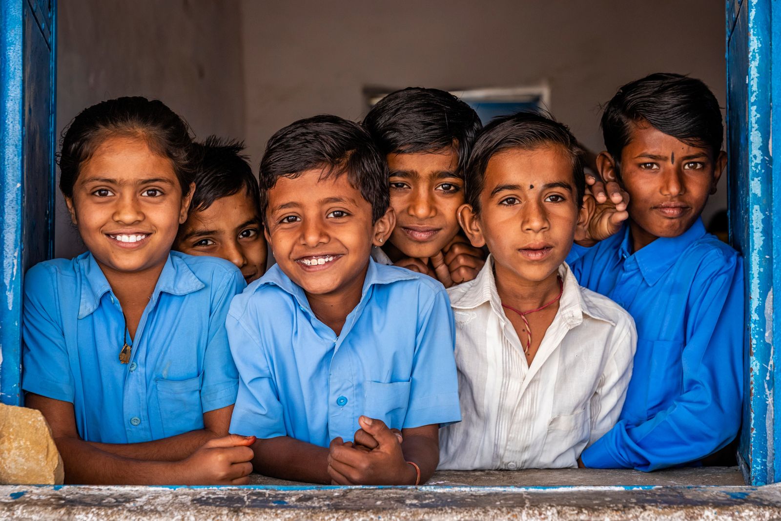 Five schoolchildren in blue uniforms smiling through a classroom window