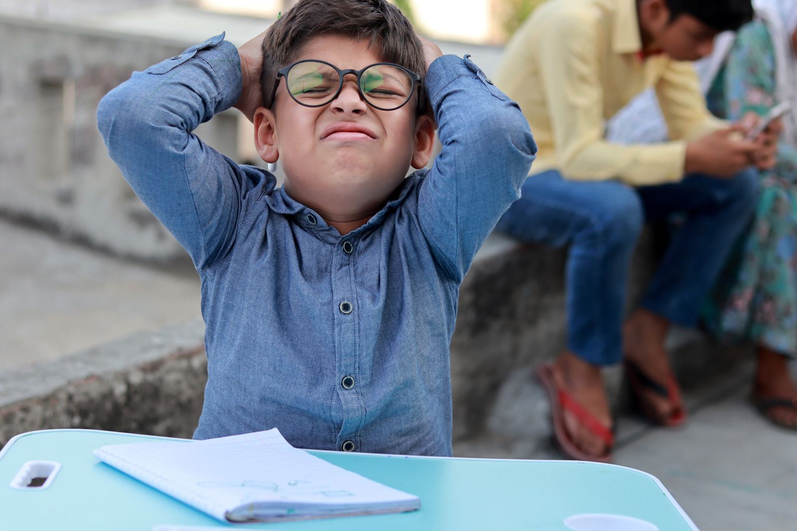 A boy in glasses with his hands on his head, thinking hard over his notebook